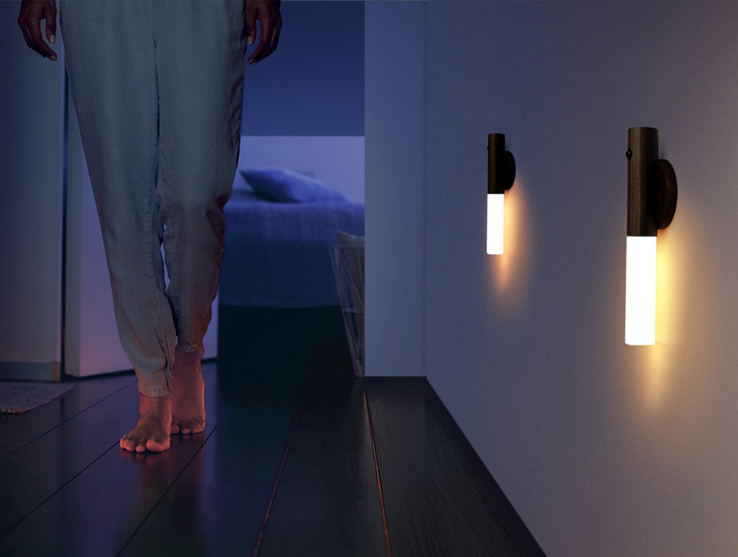 Person standing in a dimly lit room with two wall-mounted lights on a white wall.