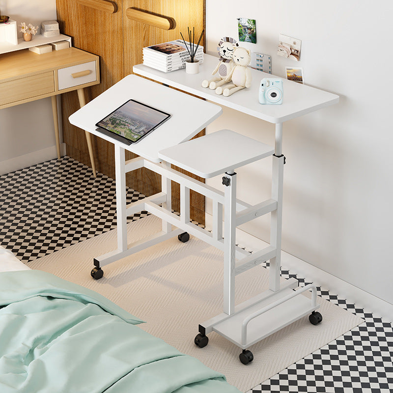 White adjustable desk with a tablet on top, placed next to a bed in a room.